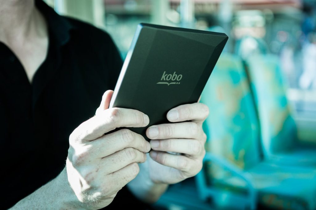 Man holding a Kobo e-reader while traveling on public transportation, focused on reading.